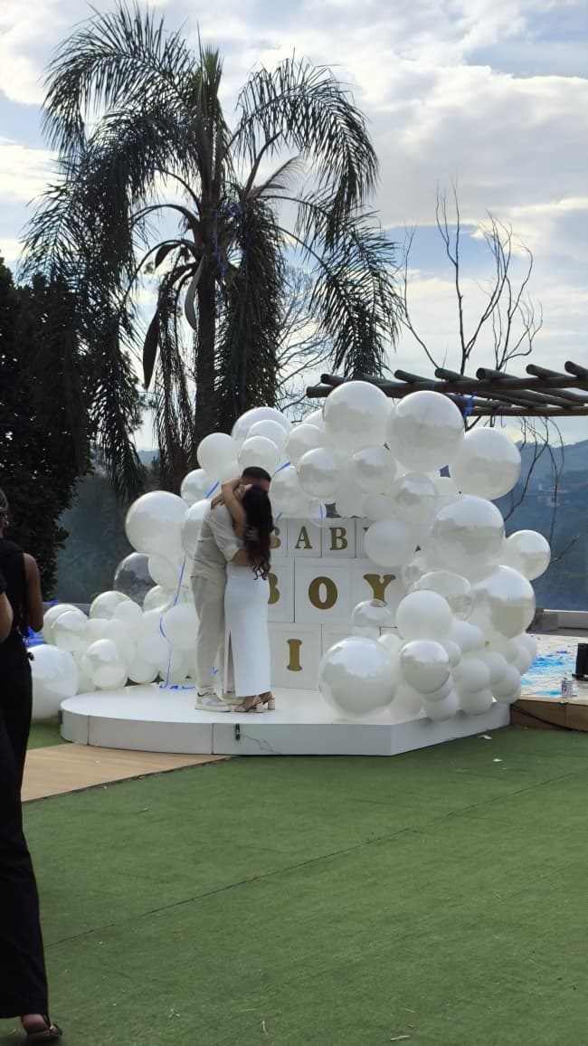 Casal diante de um arco de balões brancos com a revelação do bebê, no gramado